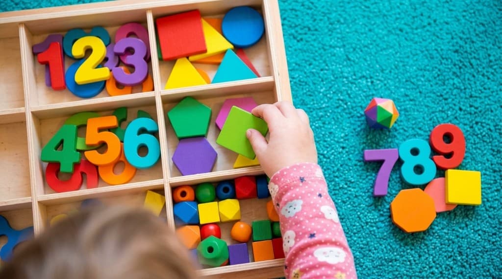 A child sorting wooden numbers and colored shapes into the compartments of a wooden tray.